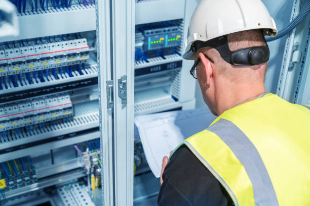 Electrician reading a wiring diagram in a control cabinet, assessing switchboard upgrades for electrical safety and efficiency.